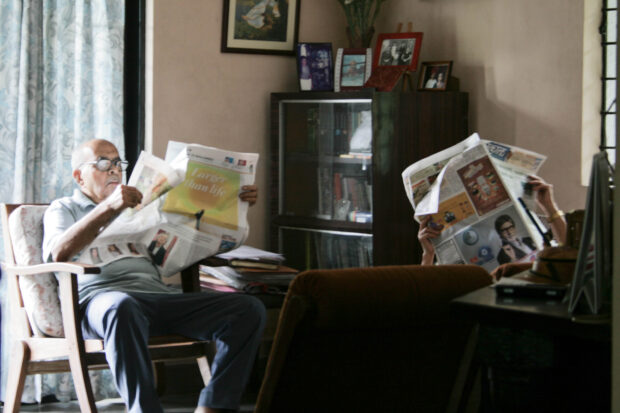 Two people in the room sitting in an arm chair reading newspapers